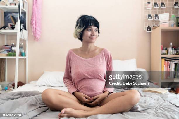 pregnant brown skin asian woman with short highlighted hair sitting on the bed at room smiling positively