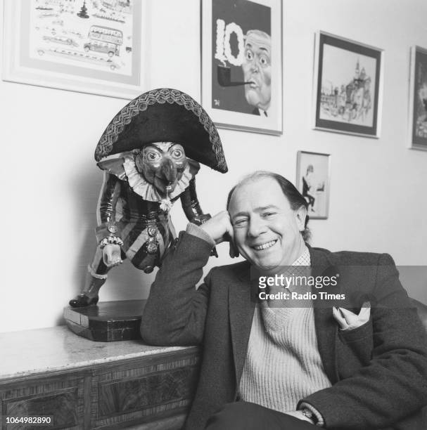 Portrait of writer and satirist Alan Coren, editor of 'Punch' magazine, sitting next to a 'Punch' character figurine, February 1983.
