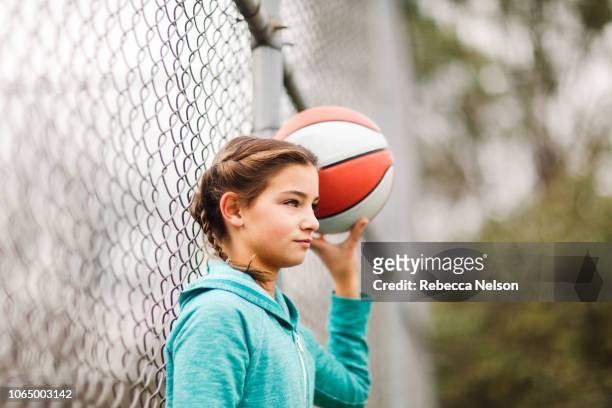 pre-teen girl leaning against chain link fence at playground, holding a basketball - chain fence stock-fotos und bilder