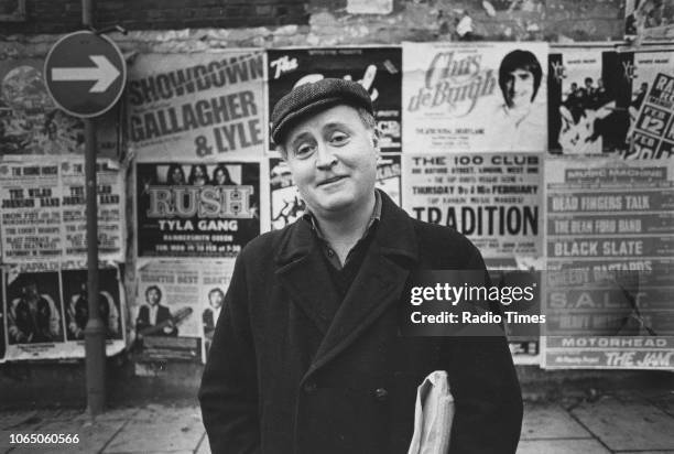 Portrait of playwright Edward Bond standing in front of a wall of posters, photographed for Radio Times in connection with the television drama 'Play...