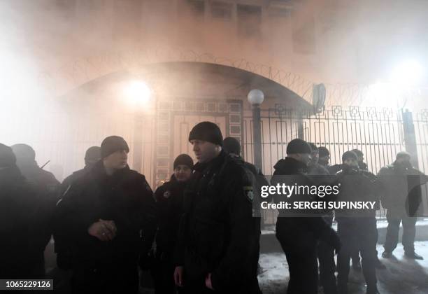 Police officers stand in front of the Russian Embassy in Kiev late on November 25 while smoke from a flare thrown by protesters is seen during a...