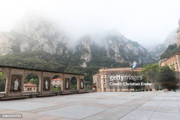 Abbey Montserrat at the sqaure in front of it with view at Roca de St. Jaume covered in fog and clouds and some historical and more modern buildings...