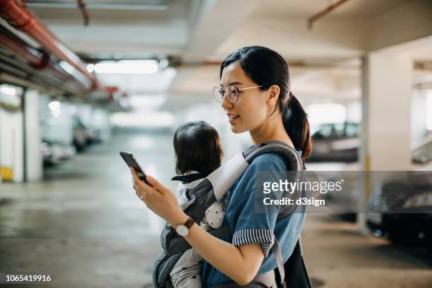 busy young woman with baby in carrier using smartphone while walking to her car in car park - madre ama de casa fotografías e imágenes de stock