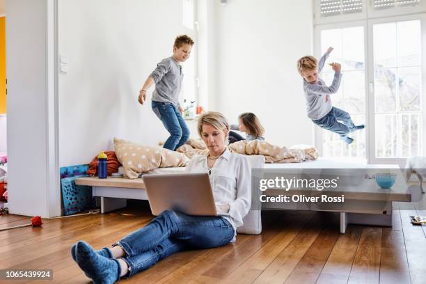 mother using laptop at home with children romping around in background - child labor fotografías e imágenes de stock