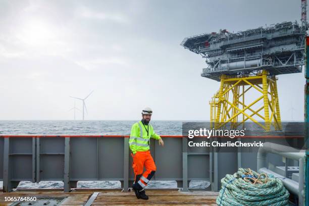 offshore manual worker standing on boat deck with platform and wind-turbines behind him - sun deck stock pictures, royalty-free photos & images