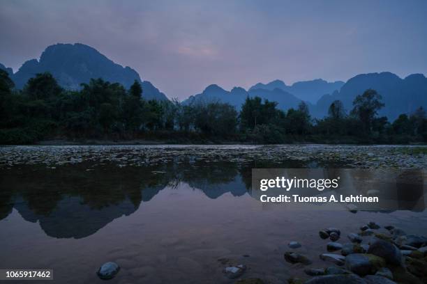 silhouette of a karst limestone mountains and their reflections on the nam song river in vang vieng, vientiane province, laos, at dusk. - shallow 2018 song stock pictures, royalty-free photos & images