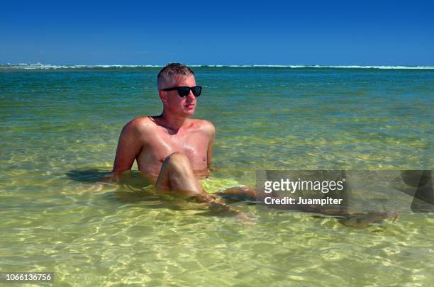 hombre joven sentado en una laguna poco profunda disfruta del sol y la playa - hombre sentado stock-fotos und bilder