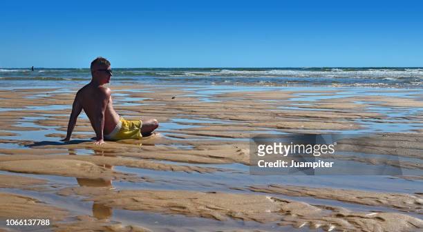 hombre joven sentado en la playa mirando al mar - hombre sentado stock-fotos und bilder