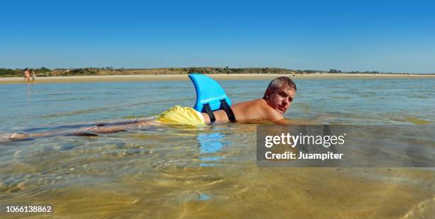 hombre joven disfruta de un baño con una aleta de tiburon en su espaldaen una laguna poco profunda disfrutando del sol y la playa - en la playa ストックフォトと画像