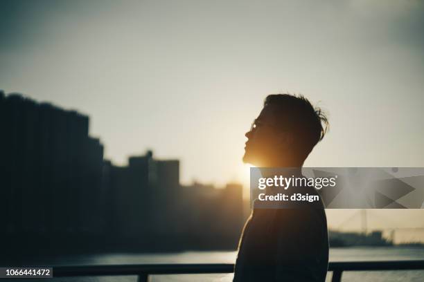 silhouette of young man standing against urban cityscape and harbour looking up to sky in deep thought at sunset - silhouette people back lit stock pictures, royalty-free photos & images