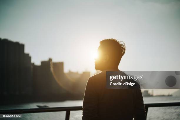 silhouette of young man standing against urban cityscape and harbour looking up to sky in deep thought - contraluz - fotografias e filmes do acervo