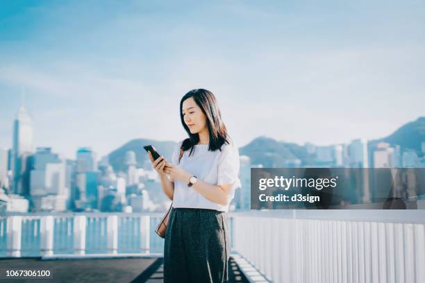 confident businesswoman checking smartphone on urban balcony against prosperous hong kong city skyline - asia skyline stock pictures, royalty-free photos & images