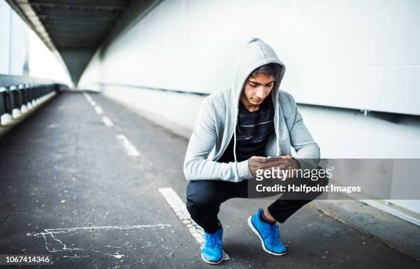 young sporty man using smartphone under the bridge in the city. - hood clothing stock pictures, royalty-free photos & images
