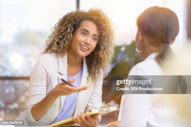 uma fêmea jovem detém uma prancheta e gestos para outra mulher enquanto está sentado em um escritório - profissional de saúde mental - fotografias e filmes do acervo