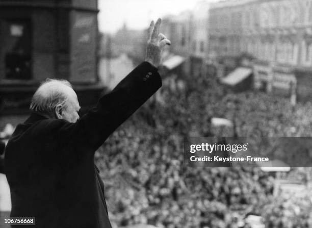British Prime Minister Winston Churchill gives his famous victory sign from the balcony of the Ministry of Health building to a huge crowd below as...
