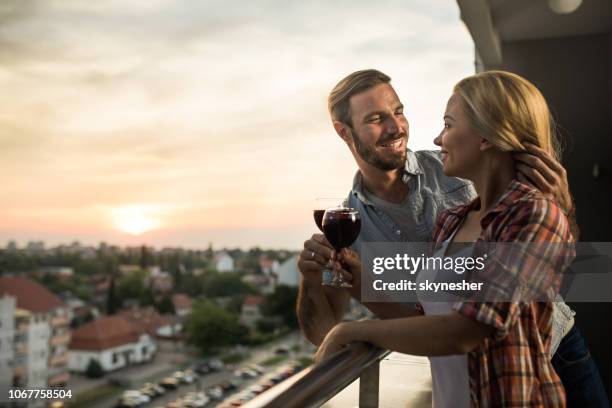 happy couple in love talking while toasting with wine on a penthouse balcony at sunset. - penthouse stock pictures, royalty-free photos & images