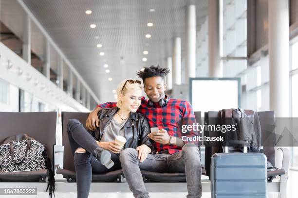 young funky couple using mobile phone in airport lounge - couple airport stock pictures, royalty-free photos & images