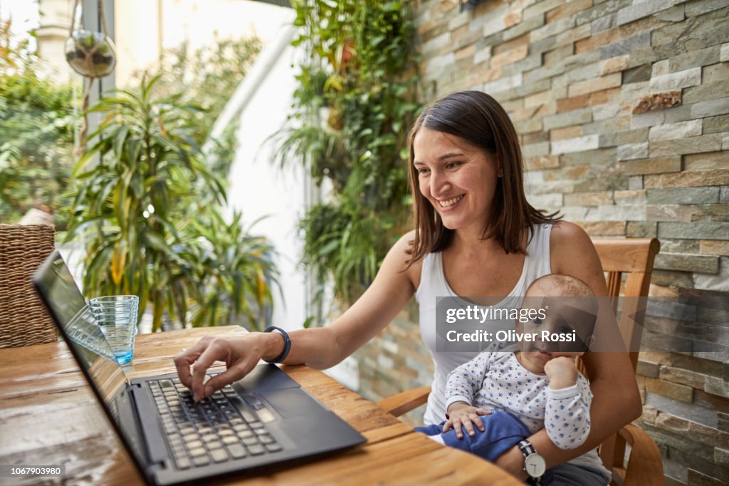 Smiling mother holding baby son and using laptop at table