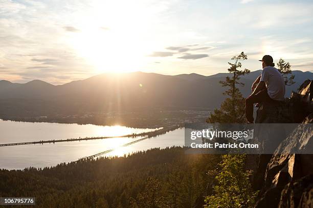 young man sits on the edge of a cliff watching the sunsset over a lake. - pend orielle lake stock pictures, royalty-free photos & images