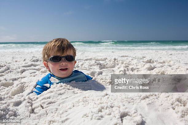 a little boy with sunglasses is buried in the sand up to his head with blue sky and ocean in the background. - destin beach stock-fotos und bilder