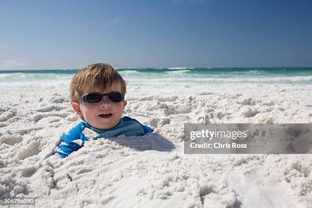 a little boy with sunglasses is buried in the sand up to his head with blue sky and ocean in the background. - destin stock pictures, royalty-free photos & images