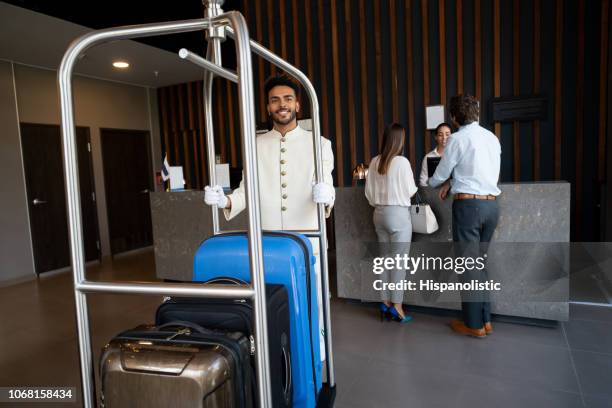 portrait of black young bellhop pushing luggage cart and business couple checking in at background - bell boy stock pictures, royalty-free photos & images