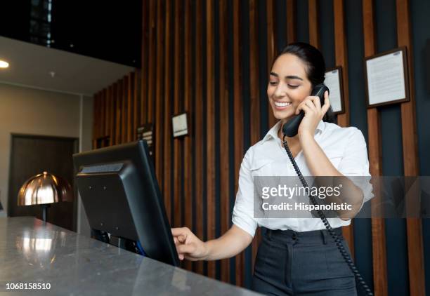 young latin american receptionist logging a booking into the system while talking on phone smiling - answering stock pictures, royalty-free photos & images