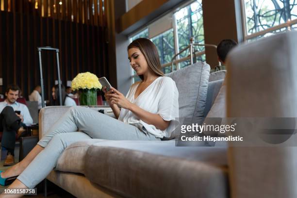beautiful latin american woman checking her smartphone while waiting at hotel lounge smiling - man waiting couch stock pictures, royalty-free photos & images
