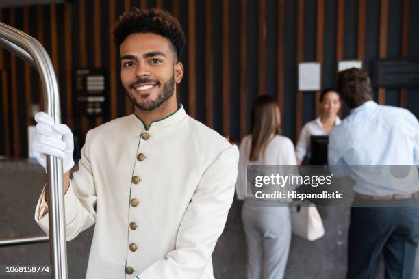 handsome young black bellhop looking at camera smiling while holding luggage cart - bell hop stock pictures, royalty-free photos & images