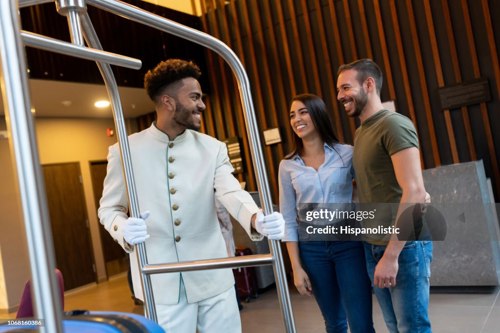 Friendly bellhop welcoming couple at hotel taking their luggage on cart all smiling