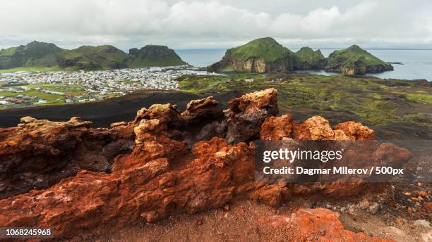 landscape with red rocks in front of town near coastline - westman islands stock pictures, royalty-free photos & images