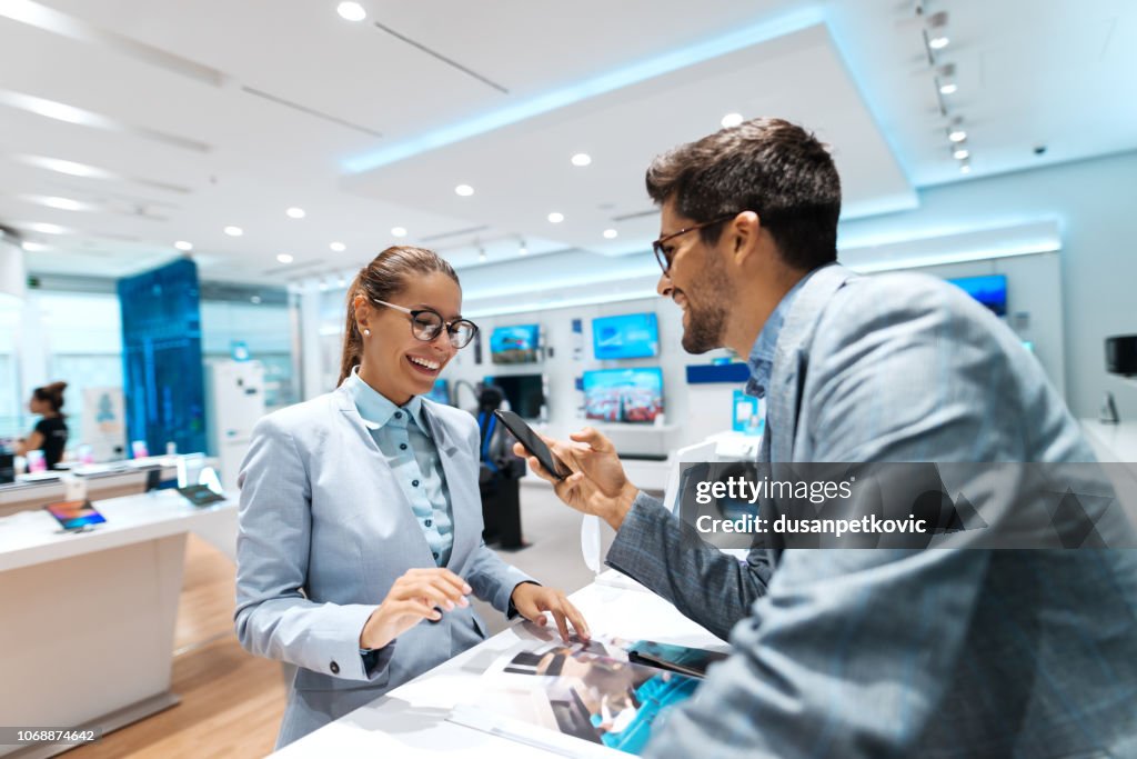 Young multicultural couple looking for new smart phone to buy. Woman looking at catalog while man holding smart phone.