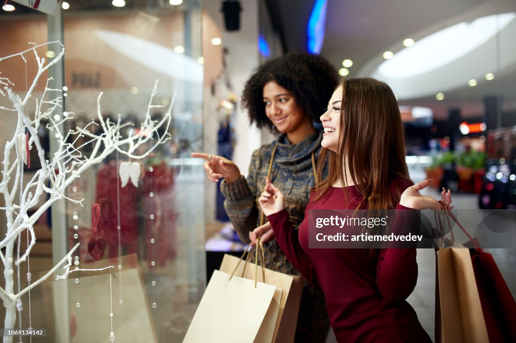 Giovani donne multietniche diverse da corsa con borse della spesa di carta vicino a vetrine decorate. Ragazza puntata a dito contro vetrina, vestiti e prodotti nel centro commerciale durante la vendita del capodanno natalizio.