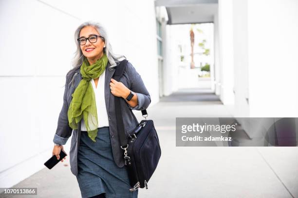 senior hispanic businesswoman walking down hallway - baby boomer stock pictures, royalty-free photos & images