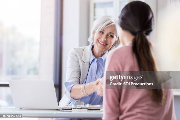 a senior woman and young woman shake hands and smile over a table - secret handshake stock pictures, royalty-free photos & images