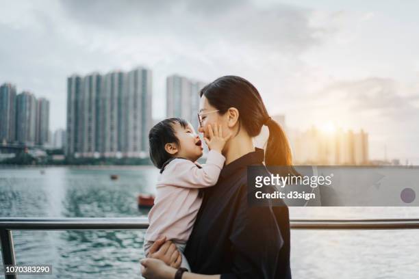 young asian mother embracing and kissing little daughter by the harbour in city at beautiful sunset - embrasser sur la bouche photos et images de collection