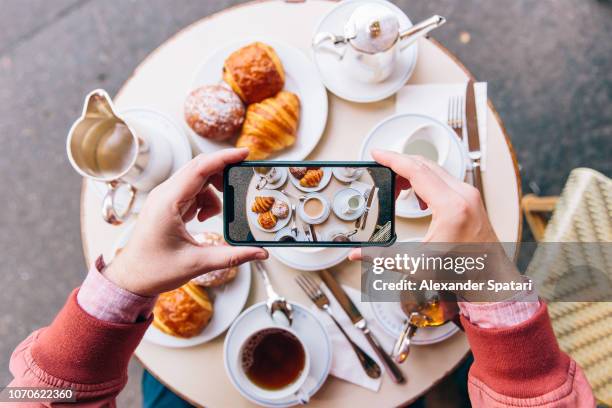 young man photographing french breakfast with croissants on the table in sidewalk cafe with smartphone, paris, france - bakery display stock-fotos und bilder