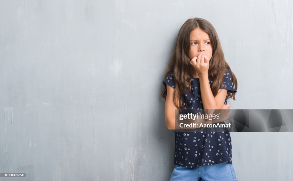 Young hispanic kid over grunge grey wall looking stressed and nervous with hands on mouth biting nails. Anxiety problem.