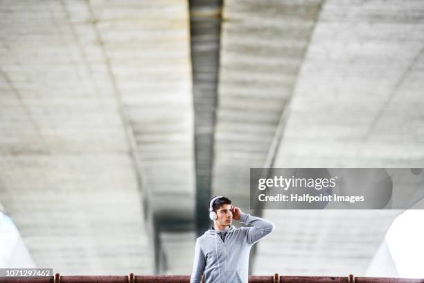 young sporty man with headphones standing under the bridge in the city, listening to music. - hoodie headphones - fotografias e filmes do acervo