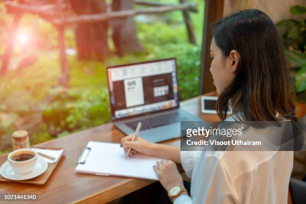 asia women are sitting thinking and writing with pen and open notebook in cafe - open laptop on desk stock-fotos und bilder