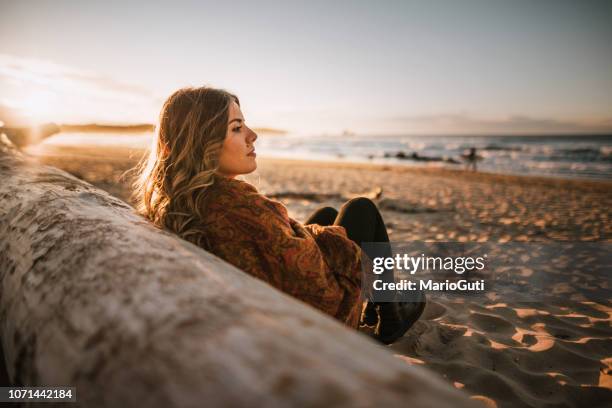 jeune femme assise sur une plage au coucher du soleil en hiver - femme rêveuse photos et images de collection