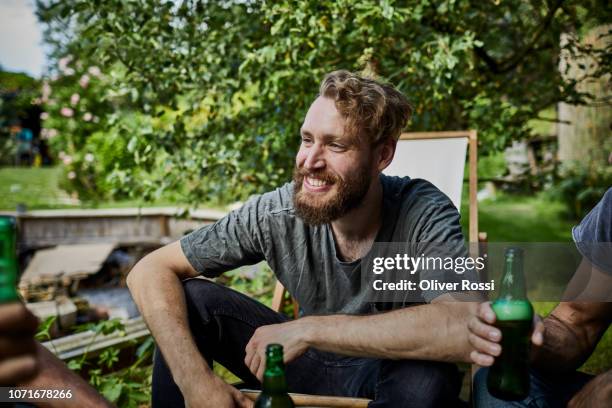 portrait of smiling man with beer bottle sitting together with friends in garden - man sipping beer smiling stockfoto's en -beelden