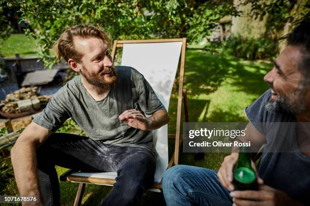 two happy men sitting in garden talking and drinking beer - man sipping beer smiling stockfoto's en -beelden