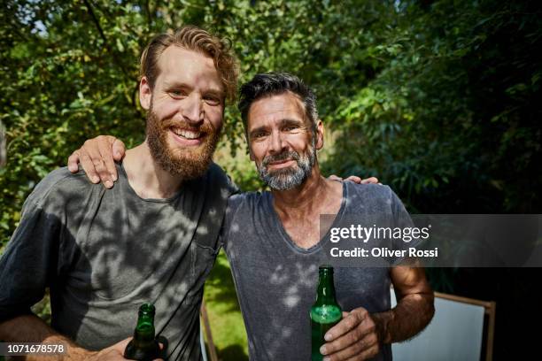 portrait of two smiling man with beer bottles embracing in garden - man sipping beer smiling stockfoto's en -beelden