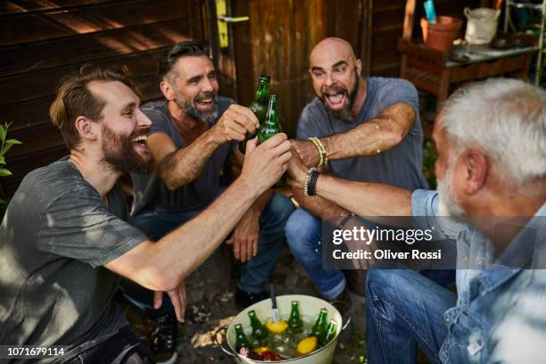 cheerful friends sitting at garden shed clinking beer bottles - bier anstoßen stock-fotos und bilder