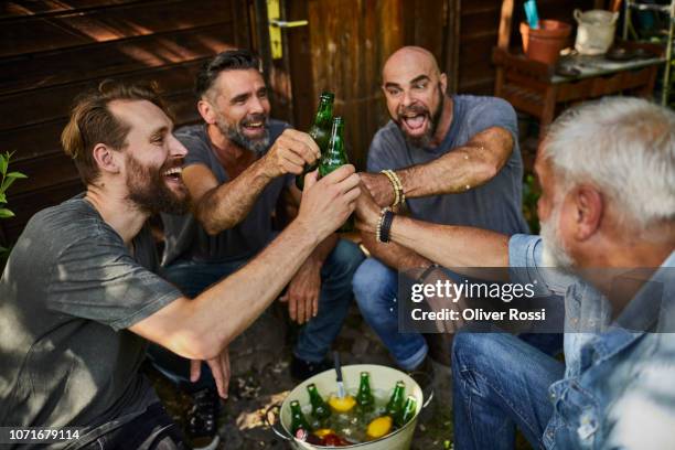 cheerful friends sitting at garden shed clinking beer bottles - nur männer stock-fotos und bilder