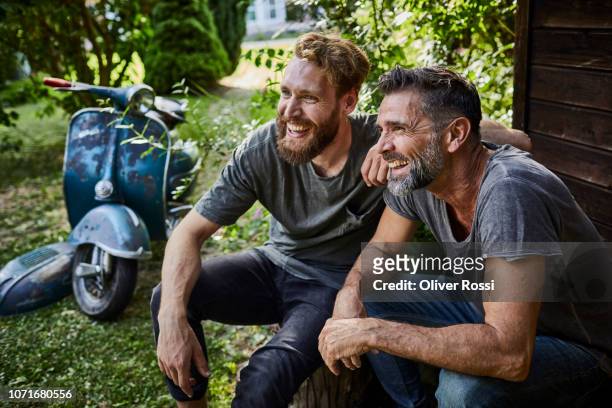 two happy men sitting together at garden shed with old motor scooter in background - mid adult men stock pictures, royalty-free photos & images