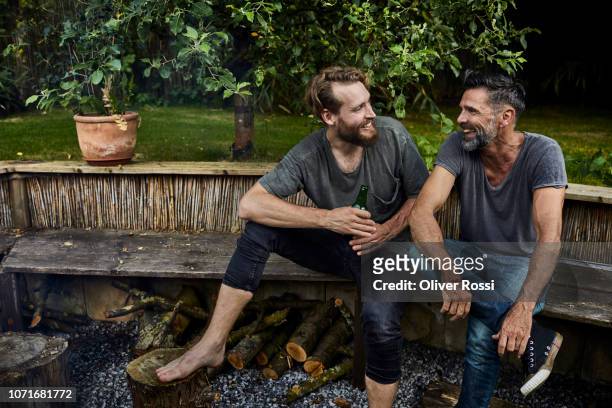 two happy men sitting together on bench in garden talking and drinking beer - man on bench stock-fotos und bilder