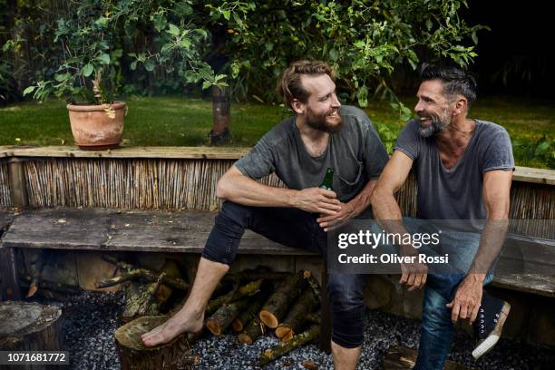 two happy men sitting together on bench in garden talking and drinking beer - garden bench stock pictures, royalty-free photos & images