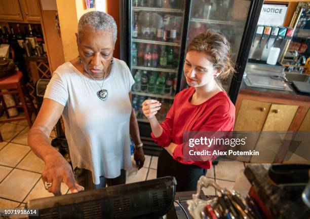 the active senior, 77-years-old, african-american businesswoman, business owner, teaching the new employee, the 18-years-old caucasian white girl, how to use the computerized cash register in the small local restaurant. - 18 19 years stock pictures, royalty-free photos & images